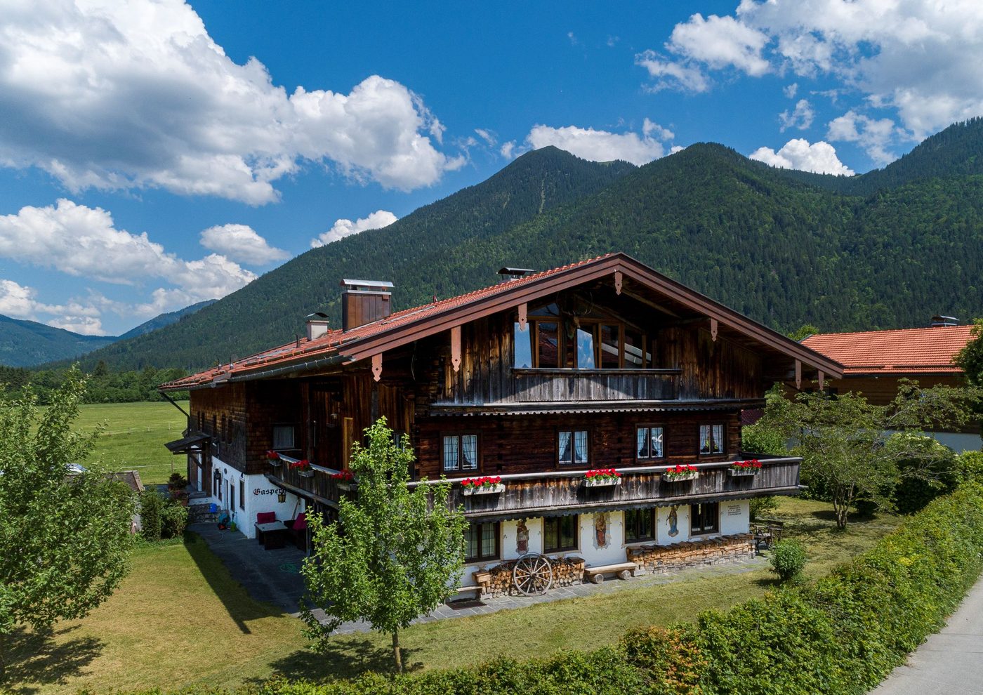 Der Gasperhof von Südwesten: dunkles Holzhaus mit weißem Erdgeschoss und Geranien an der Laube, dahinter bewaldete Berge